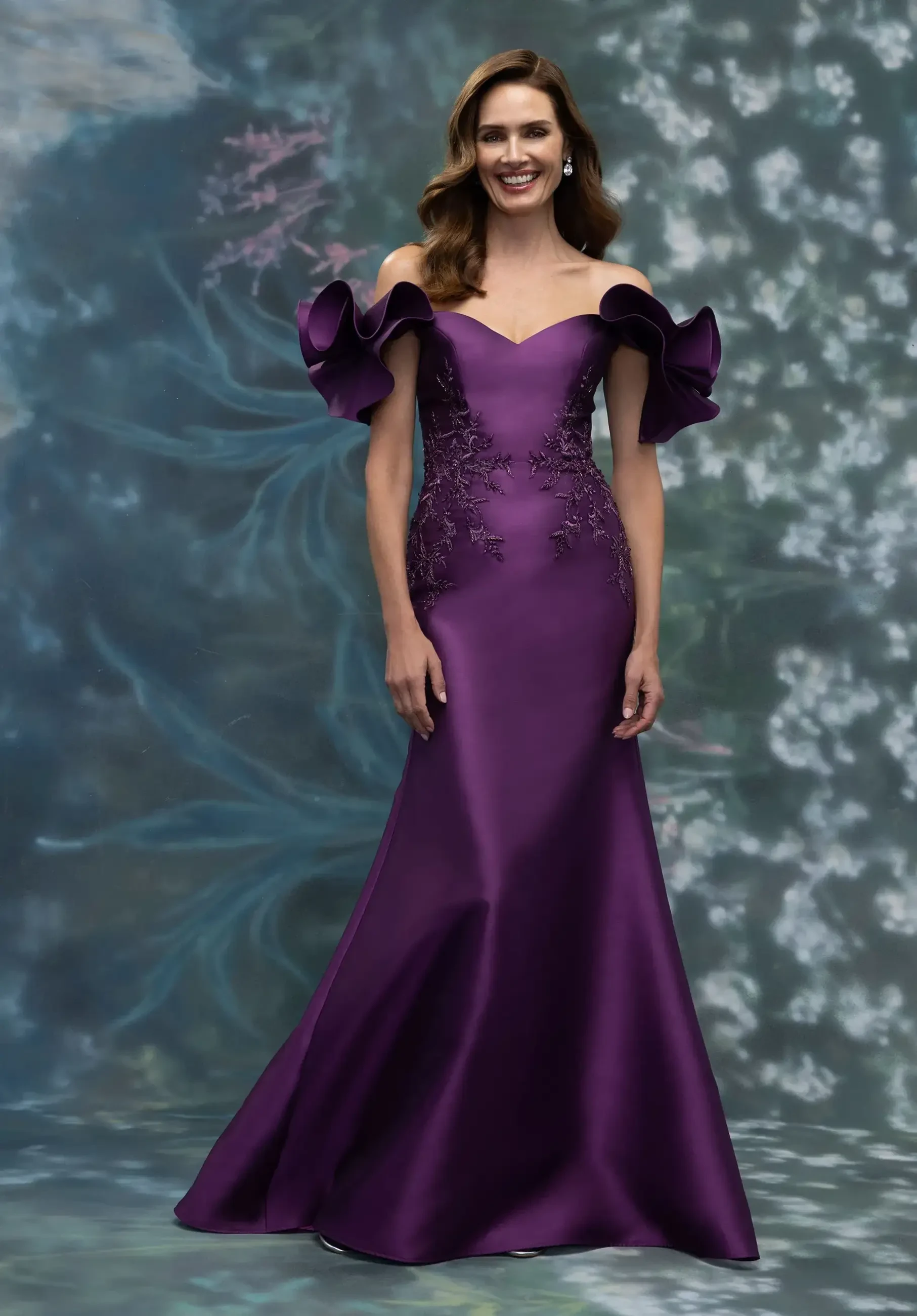 A woman wearing an off-shoulder purple gown with ruffled sleeves and embroidered details, standing in front of a floral backdrop.