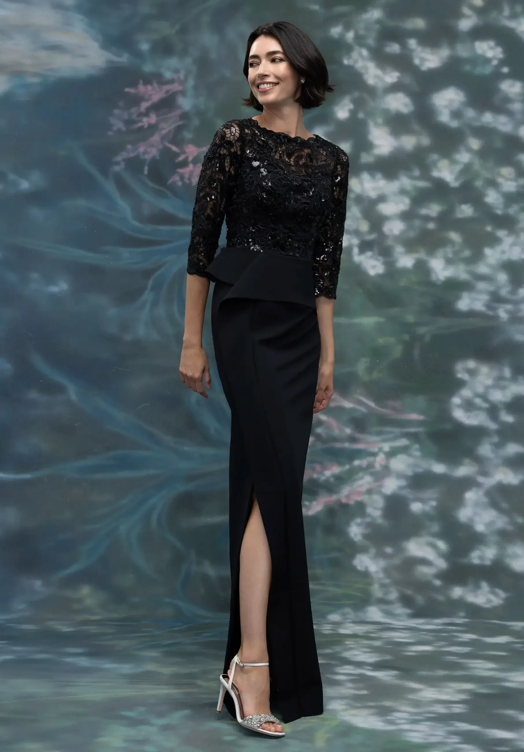 A woman wearing an elegant black dress with a lace top and a fitted skirt featuring a thigh-high slit, standing against a soft floral background.