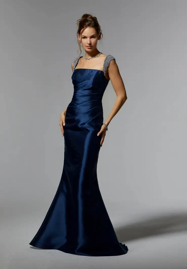 A woman wearing a sleek, navy blue evening gown with embellished shoulder straps, posing confidently against a neutral background.