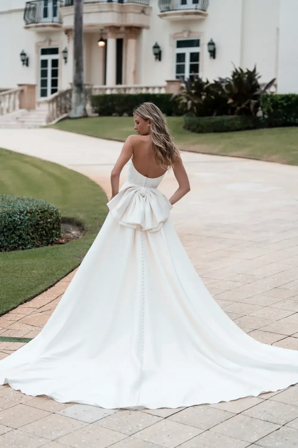 A woman in a white gown stands outdoors, facing away from the camera. The gown has a dramatic train and a bow detail at the back. She is on a paved pathway in a landscaped area.