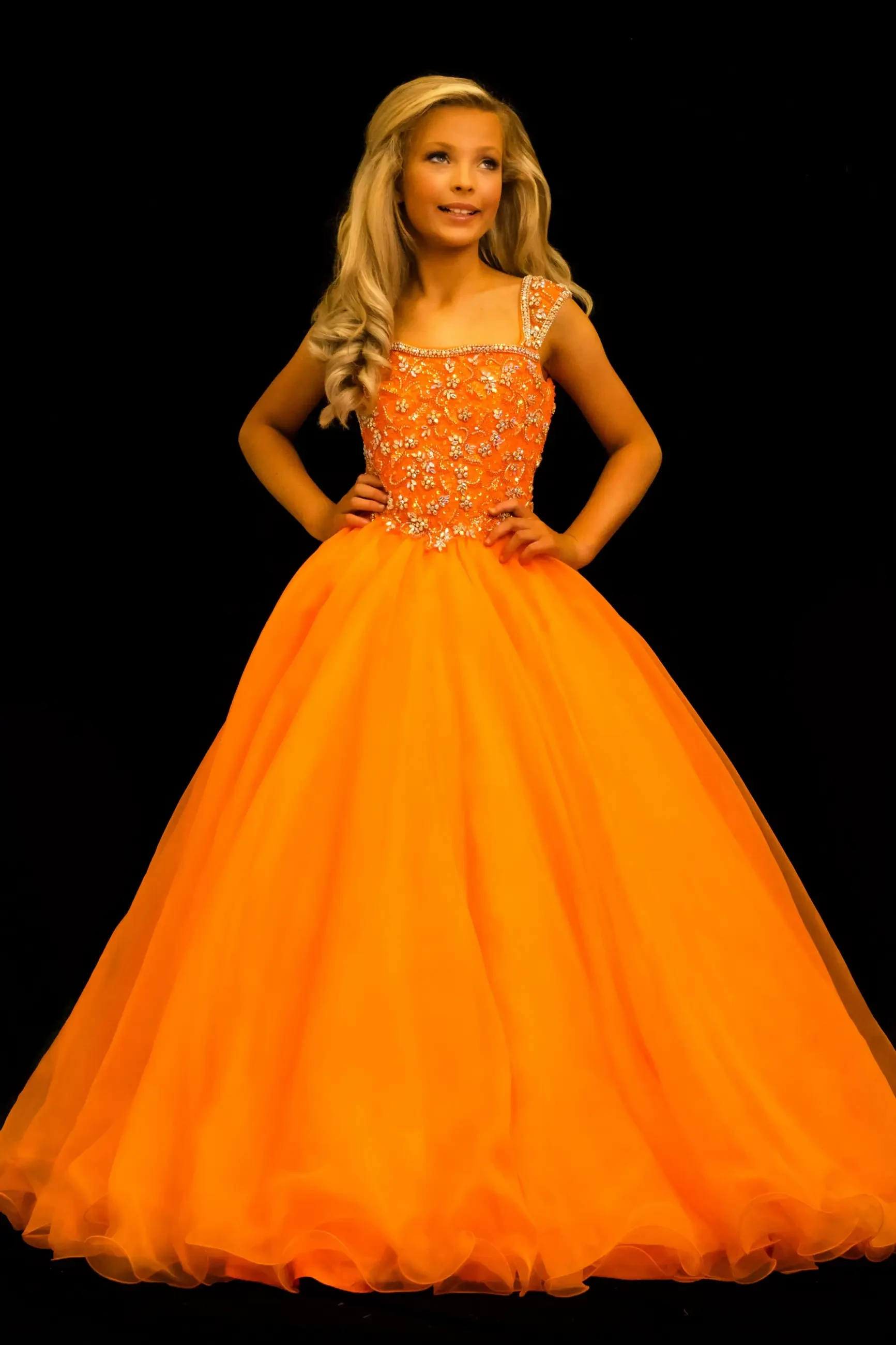 A young girl wearing an elegant orange ball gown with decorative embellishments, standing confidently against a black background.