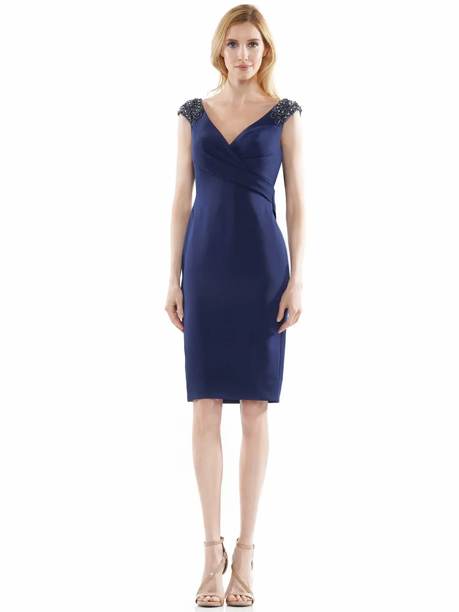 A woman stands wearing a navy blue dress with short embellished sleeves and a fitted silhouette, paired with strappy heels.