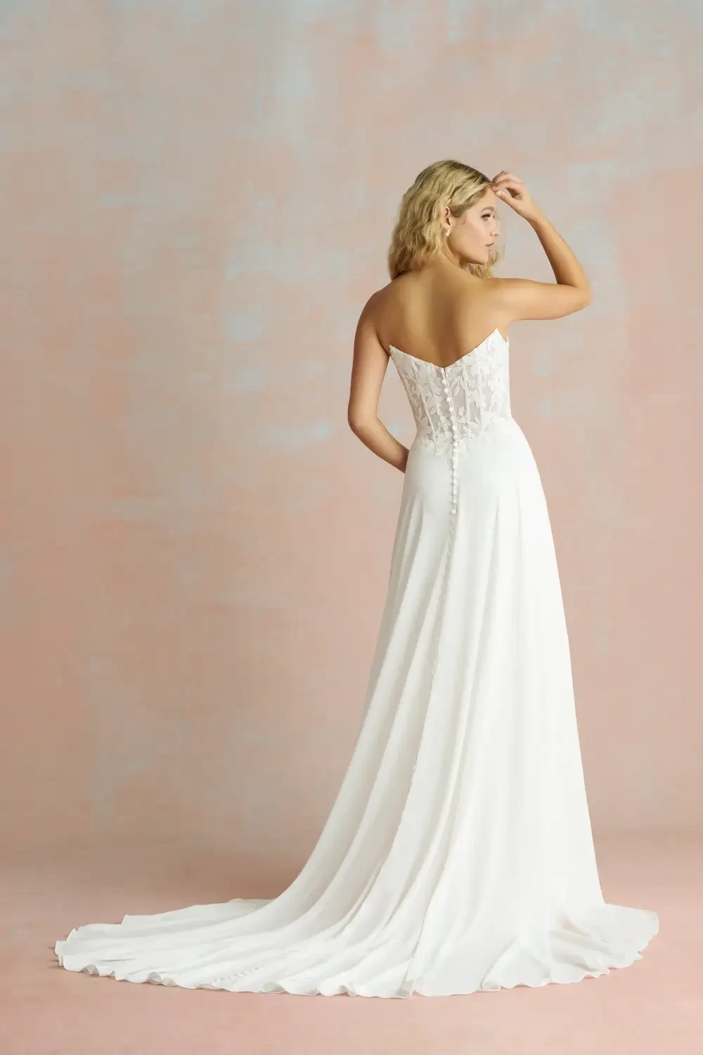 A model wearing a white gown with a fitted bodice and a flowing skirt, posed from the back against a pink textured background.